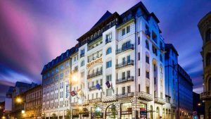 Una vista de la calle del gran y ornamentado hotel Novotel Budapest Centrum al anochecer, con luces brillantes que proyectan un resplandor romántico bajo el cielo púrpura de Budapest. - ForTwo - Planes para parejas