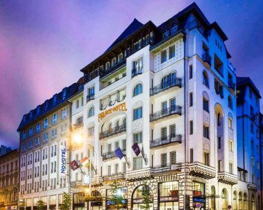 Una vista de la calle del gran y ornamentado hotel Novotel Budapest Centrum al anochecer, con luces brillantes que proyectan un resplandor romántico bajo el cielo púrpura de Budapest. - ForTwo - Planes para parejas
