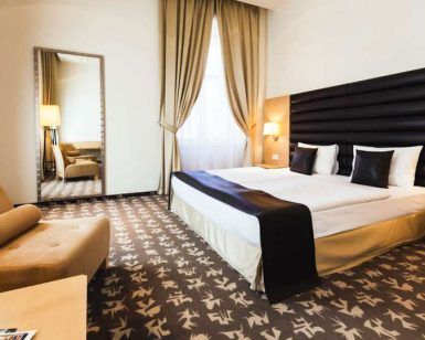 El Hotel Buda Castle Fashion, en el centro de Budapest, ofrece una habitación moderna con cama doble, sillón, espejo grande, alfombra texturizada e iluminación cálida. - ForTwo - Planes para parejas