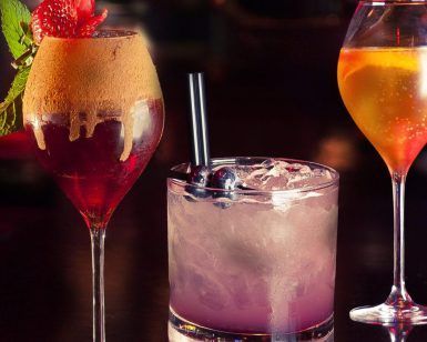 Experimente la rica coctelería de Barcelona con Taller de Coctelería: Aprende a Preparar y Degustar 3 Cócteles Exclusivos, que presenta un elegante cóctel con guarnición de fresas, uno con arándanos frescos y una pajita, y una obra maestra en tonos naranjas. - ForTwo - Planes para parejas