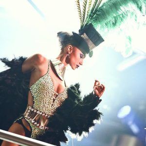 Una artista con un traje brillante y un sombrero de plumas verdes adopta una pose dramática en el escenario de SHÔKO Barcelona, todo ello bajo el resplandor de un foco. - ForTwo - Planes para parejas