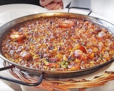 Una sartén con la tradicional paella de marisco, gambas y verduras, que recuerda a los sabores del Restaurante 7 Portes (Barcelona), reposa elegantemente sobre una cesta tejida. - ForTwo - Planes para parejas