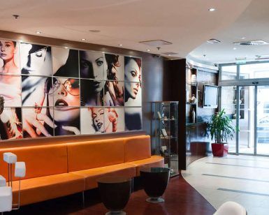El paquete Escapadita a Budapest en pareja con hotel y desayuno incluye una estadía en un moderno lobby de hotel adornado con asientos naranjas, vibrantes retratos de arte en las paredes, exuberantes plantas y una elegante entrada de vidrio para un ambiente acogedor. - ForTwo - Planes para parejas