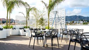 Un patio en la azotea con sillas negras, sofás blancos y palmeras ofrece el entorno perfecto para una escapada romántica en un hotel con jacuzzi y cena en Torremolinos. Relájese junto a la celosía decorativa en este tranquilo oasis bajo un cielo parcialmente nublado. - ForTwo - Planes para parejas