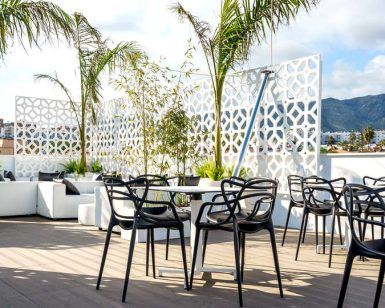 Un patio en la azotea con sillas negras, sofás blancos y palmeras ofrece el entorno perfecto para una escapada romántica en un hotel con jacuzzi y cena en Torremolinos. Relájese junto a la celosía decorativa en este tranquilo oasis bajo un cielo parcialmente nublado. - ForTwo - Planes para parejas