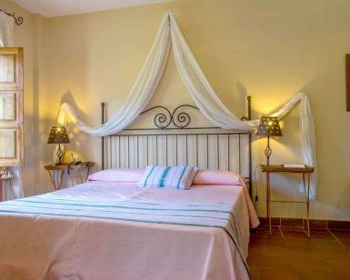 Disfrute de un acogedor refugio en el Hotel Humaina, con una colcha rosa, un cabecero ornamentado, mesitas de noche, lámparas y vistas a la montaña. Perfecto para una escapada a los Montes de Málaga, con un cuadro decorativo como toque final. - ForTwo - Planes para parejas