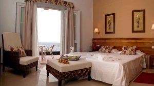 Disfrute de la Escapada en El Hierro con spa en el Hotel Balneario Pozo de la Salud. Su habitación ofrece una cómoda cama doble, una silla de mimbre y un balcón con vistas al mar. Disfrute de una decoración serena con cojines florales y obras de arte enmarcadas para un remanso de paz. - ForTwo - Planes para parejas