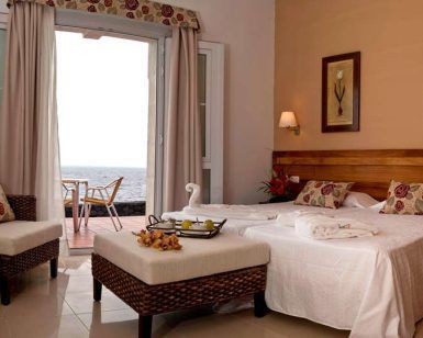 Disfrute de la Escapada en El Hierro con spa en el Hotel Balneario Pozo de la Salud. Su habitación ofrece una cómoda cama doble, una silla de mimbre y un balcón con vistas al mar. Disfrute de una decoración serena con cojines florales y obras de arte enmarcadas para un remanso de paz. - ForTwo - Planes para parejas