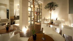 La moderna cafetería interior del Hotel Taburiente en Tenerife ofrece sillas blancas, mesas de madera, exuberantes plantas en macetas y una cálida iluminación. Grandes puertas de cristal dan la bienvenida a la luz natural en el corazón de la ciudad. - ForTwo - Planes para parejas