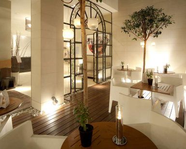 La moderna cafetería interior del Hotel Taburiente en Tenerife ofrece sillas blancas, mesas de madera, exuberantes plantas en macetas y una cálida iluminación. Grandes puertas de cristal dan la bienvenida a la luz natural en el corazón de la ciudad. - ForTwo - Planes para parejas