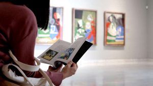 En el Museo Picasso Málaga, una persona se pierde en un libro, rodeada de pinturas abstractas, lo que proporciona un telón de fondo ideal para una escapada romántica llena de arte con la Escapada romántica a Málaga con entradas al museo Picasso: hotel María Cristina. - ForTwo - Planes para parejas