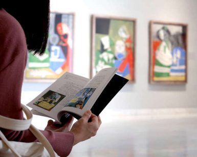 En el Museo Picasso Málaga, una persona se pierde en un libro, rodeada de pinturas abstractas, lo que proporciona un telón de fondo ideal para una escapada romántica llena de arte con la Escapada romántica a Málaga con entradas al museo Picasso: hotel María Cristina. - ForTwo - Planes para parejas