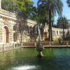 Bajo un cielo azul despejado, el ornamentado edificio de piedra con una fuente junto a un estanque rodeado de palmeras evoca la exuberante vegetación de Sevilla. Experimente esta magia en el Real Alcázar con "Visita el Real Alcázar de Sevilla sin esperas" para una aventura inolvidable. - ForTwo - Planes para parejas