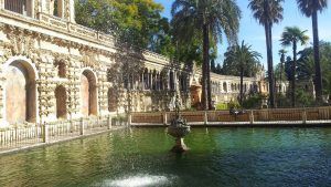 Bajo un cielo azul despejado, el ornamentado edificio de piedra con una fuente junto a un estanque rodeado de palmeras evoca la exuberante vegetación de Sevilla. Experimente esta magia en el Real Alcázar con "Visita el Real Alcázar de Sevilla sin esperas" para una aventura inolvidable. - ForTwo - Planes para parejas