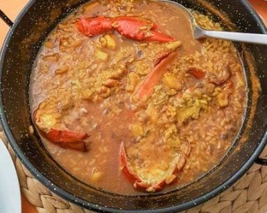 Una paella de mariscos con pimientos rojos y una cuchara para servir reposa sobre un salvamanteles tejido, capturando los vibrantes sabores del Restaurante Arrozeando (Málaga). Perfecto para disfrutar en este encantador restaurante malagueño. - ForTwo - Planes para parejas
