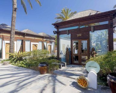 La oferta de spa, yoga y almuerzo en Atzaro (Ibiza) ofrece un entorno sereno con decoración de madera, plantas en macetas, cestas de frutas y palmeras bajo un cielo azul claro, perfecto para sesiones de yoga. - ForTwo - Planes para parejas