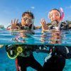 Dos buceadores con sus equipos flotan sobre las cristalinas aguas de Tarifa, haciendo señas con las manos. El bautismo de buceo con goDive en Tarifa (Cádiz) ofrece una introducción inolvidable, mientras barcos y edificios crean un pintoresco escenario para esta aventura. - ForTwo - Planes para parejas