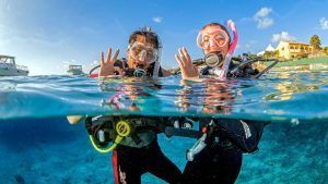 Dos buceadores con sus equipos flotan sobre las cristalinas aguas de Tarifa, haciendo señas con las manos. El bautismo de buceo con goDive en Tarifa (Cádiz) ofrece una introducción inolvidable, mientras barcos y edificios crean un pintoresco escenario para esta aventura. - ForTwo - Planes para parejas