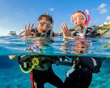 Dos buceadores con sus equipos flotan sobre las cristalinas aguas de Tarifa, haciendo señas con las manos. El bautismo de buceo con goDive en Tarifa (Cádiz) ofrece una introducción inolvidable, mientras barcos y edificios crean un pintoresco escenario para esta aventura. - ForTwo - Planes para parejas