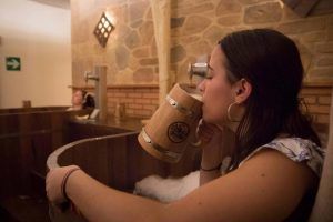 En un rústico Beer Spa de Tenerife, una mujer disfruta de una relajante bañera de madera mientras bebe de una taza también del mismo material. Otro huésped se relaja cerca, creando una experiencia idílica en el Circuito Beer Spa para dos que te transporta a la tranquilidad. - ForTwo - Planes para parejas