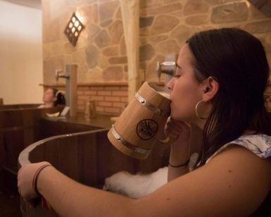 En un rústico Beer Spa de Tenerife, una mujer disfruta de una relajante bañera de madera mientras bebe de una taza también del mismo material. Otro huésped se relaja cerca, creando una experiencia idílica en el Circuito Beer Spa para dos que te transporta a la tranquilidad. - ForTwo - Planes para parejas