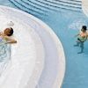 Dos personas disfrutan de un momento de serenidad en el centro de bienestar Caldea de Andorra con la experiencia "Entrada al spa de Caldea": uno se relaja en un espacio circular, mientras el otro disfruta de la relajante cascada de un chorro de agua. - ForTwo - Planes para parejas