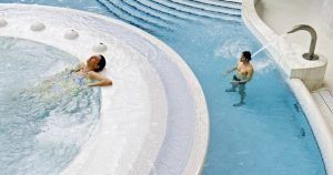 Dos personas disfrutan de un momento de serenidad en el centro de bienestar Caldea de Andorra con la experiencia "Entrada al spa de Caldea": uno se relaja en un espacio circular, mientras el otro disfruta de la relajante cascada de un chorro de agua. - ForTwo - Planes para parejas
