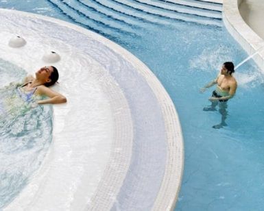 Dos personas disfrutan de un momento de serenidad en el centro de bienestar Caldea de Andorra con la experiencia "Entrada al spa de Caldea": uno se relaja en un espacio circular, mientras el otro disfruta de la relajante cascada de un chorro de agua. - ForTwo - Planes para parejas