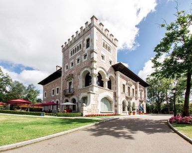 La impresionante mansión de piedra, que evoca al Hotel Castillo Del Bosque La Zoreda de Oviedo, cuenta con una torre rodeada de exuberantes jardines con un camino sinuoso bajo un cielo parcialmente nublado. - ForTwo - Planes para parejas