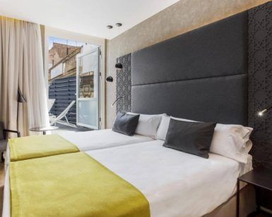 Moderna habitación con dos camas individuales en el Hotel Catalonia Giralda, con cabecero gris y manta verde. Balcón con vistas a la histórica Giralda a través de una ventana abierta con cortina. - ForTwo - Planes para parejas