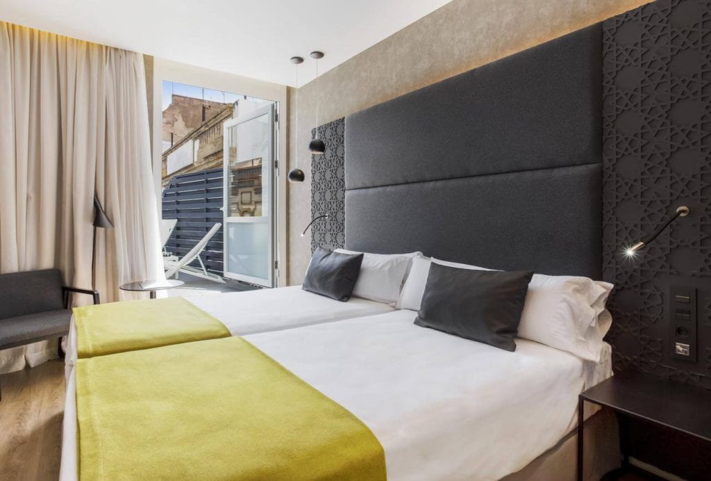 Moderna habitación con dos camas individuales en el Hotel Catalonia Giralda, con cabecero gris y manta verde. Balcón con vistas a la histórica Giralda a través de una ventana abierta con cortina. - ForTwo - Planes para parejas