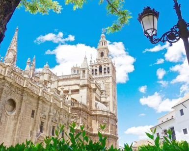 Experimente una vista impresionante de la icónica torre de la Giralda de la Catedral de Sevilla con su "Entrada para la Catedral de Sevilla y la Giralda (sin colas)", con un cielo azul como telón de fondo, una exuberante vegetación y acompañada de una encantadora farola vintage. - ForTwo - Planes para parejas