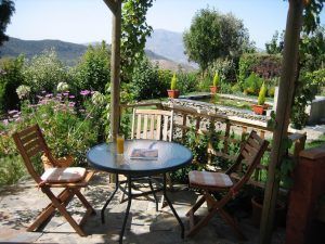 Disfrute de las pintorescas montañas de las Alpujarras desde el patio de su jardín en Apartamentos con cocina en las Alpujarras Cortijo Opazo (Granada), que cuenta con una mesa redonda de vidrio, dos sillas y flores vibrantes. - ForTwo - Planes para parejas