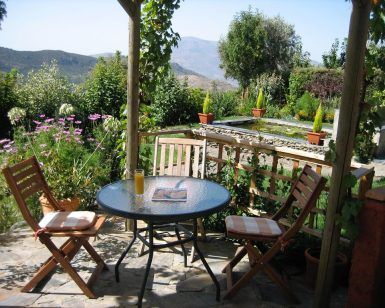 Disfrute de las pintorescas montañas de las Alpujarras desde el patio de su jardín en Apartamentos con cocina en las Alpujarras Cortijo Opazo (Granada), que cuenta con una mesa redonda de vidrio, dos sillas y flores vibrantes. - ForTwo - Planes para parejas