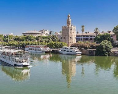 El Crucero Torre del Oro por el Guadalquivir se desliza a lo largo del tranquilo río, con la histórica Torre del Oro y pintorescos edificios en el pintoresco telón de fondo bajo un cielo azul claro. - ForTwo - Planes para parejas
