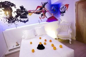 Una elegante habitación del Hotel Dormirdcine Madrid presenta una decoración blanca, un mural vibrante y detalles encantadores como naranjas y un sombrero en la cama. - ForTwo - Planes para parejas