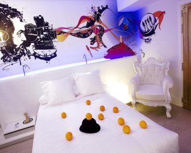 Una elegante habitación del Hotel Dormirdcine Madrid presenta una decoración blanca, un mural vibrante y detalles encantadores como naranjas y un sombrero en la cama. - ForTwo - Planes para parejas