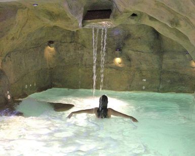 Una persona se relaja en una piscina cueva en el Cercado de la Alpujarra, con agua en cascada y una relajante iluminación ambiental, que recuerda a la experiencia del "Circuito Spa con masaje incluido para 2 personas en el Cercado de la Alpujarra". - ForTwo - Planes para parejas