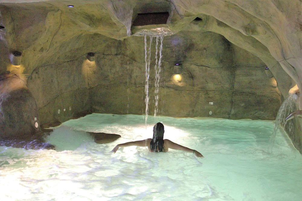 Una persona se relaja en una piscina cueva en el Cercado de la Alpujarra, con agua en cascada y una relajante iluminación ambiental, que recuerda a la experiencia del "Circuito Spa con masaje incluido para 2 personas en el Cercado de la Alpujarra". - ForTwo - Planes para parejas