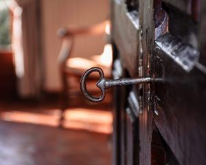 Una llave antigua en una antigua cerradura de puerta de madera evoca una sensación de misterio, similar al Escape Room Resuelve el Misterio del Hotel en Zafra, con un fondo borroso de una silla y una ventana que añade elegancia. - ForTwo - Planes para parejas