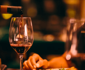 El vino tinto, rico y aterciopelado, se vierte expertamente en una copa en medio de una iluminación cálida y un fondo borroso, creando una atmósfera íntima similar a la "Excursión y Cata de Vinos de la Ribera del Duero desde Madrid". - ForTwo - Planes para parejas