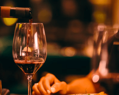 El vino tinto, rico y aterciopelado, se vierte expertamente en una copa en medio de una iluminación cálida y un fondo borroso, creando una atmósfera íntima similar a la "Excursión y Cata de Vinos de la Ribera del Duero desde Madrid". - ForTwo - Planes para parejas