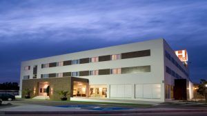 Bajo un cielo nublado por la noche, el Hotel Las Bóvedas en Badajoz impresiona con su diseño moderno y su icónico letrero H brillante. Para una experiencia inolvidable, considere el exclusivo Paquete Romántico en Hotel Las Bóvedas 4 estrellas. - ForTwo - Planes para parejas
