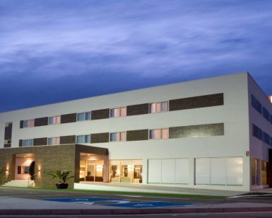 Bajo un cielo nublado por la noche, el Hotel Las Bóvedas en Badajoz impresiona con su diseño moderno y su icónico letrero H brillante. Para una experiencia inolvidable, considere el exclusivo Paquete Romántico en Hotel Las Bóvedas 4 estrellas. - ForTwo - Planes para parejas
