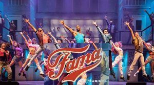 Artistas con trajes vibrantes saltan al escenario del Teatro EDP Gran Vía, con el texto "Fama, el musical" superpuesto. - ForTwo - Planes para parejas