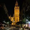 Una torre gótica brillantemente iluminada en Sevilla, rodeada de árboles y gente en calles adoquinadas, susurros de antiguas leyendas, que recuerdan a un "Tour gratuito de los misterios y leyendas de Sevilla", explorando la historia en cada sombra. - ForTwo - Planes para parejas