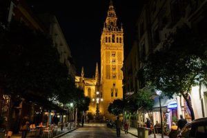 Una torre gótica brillantemente iluminada en Sevilla, rodeada de árboles y gente en calles adoquinadas, susurros de antiguas leyendas, que recuerdan a un "Tour gratuito de los misterios y leyendas de Sevilla", explorando la historia en cada sombra. - ForTwo - Planes para parejas