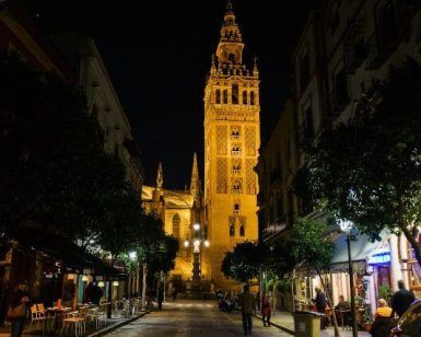 Una torre gótica brillantemente iluminada en Sevilla, rodeada de árboles y gente en calles adoquinadas, susurros de antiguas leyendas, que recuerdan a un "Tour gratuito de los misterios y leyendas de Sevilla", explorando la historia en cada sombra. - ForTwo - Planes para parejas