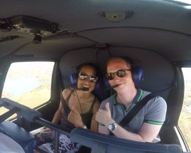 Dos personas con auriculares hacen el gesto de aprobación con el pulgar en la cabina de un helicóptero, disfrutando de un "Vuelo en helicóptero por la Costa del Sol" sobre el pintoresco paisaje. - ForTwo - Planes para parejas