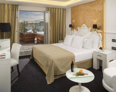 Disfrute de la elegancia del Hotel Gran Meliá Colón Sevilla con una lujosa cama, vista al balcón y una acogedora sala de estar con una mesa decorada con vino y naranjas frescas para mejorar su estadía. - ForTwo - Planes para parejas